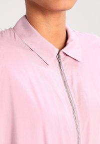 Hellrosa, mit Reißverschluss versehenes Jackett mit Kragen. Hergestellt aus glattem, leichtem Stoff. Verfügt über einen silbernen Reißverschluss und ein einfaches, minimalistisches Design.