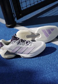 Chaussures de tennis blanches et violet clair avec des semelles texturées reposant sur un court de tennis bleu près du poteau de filet.