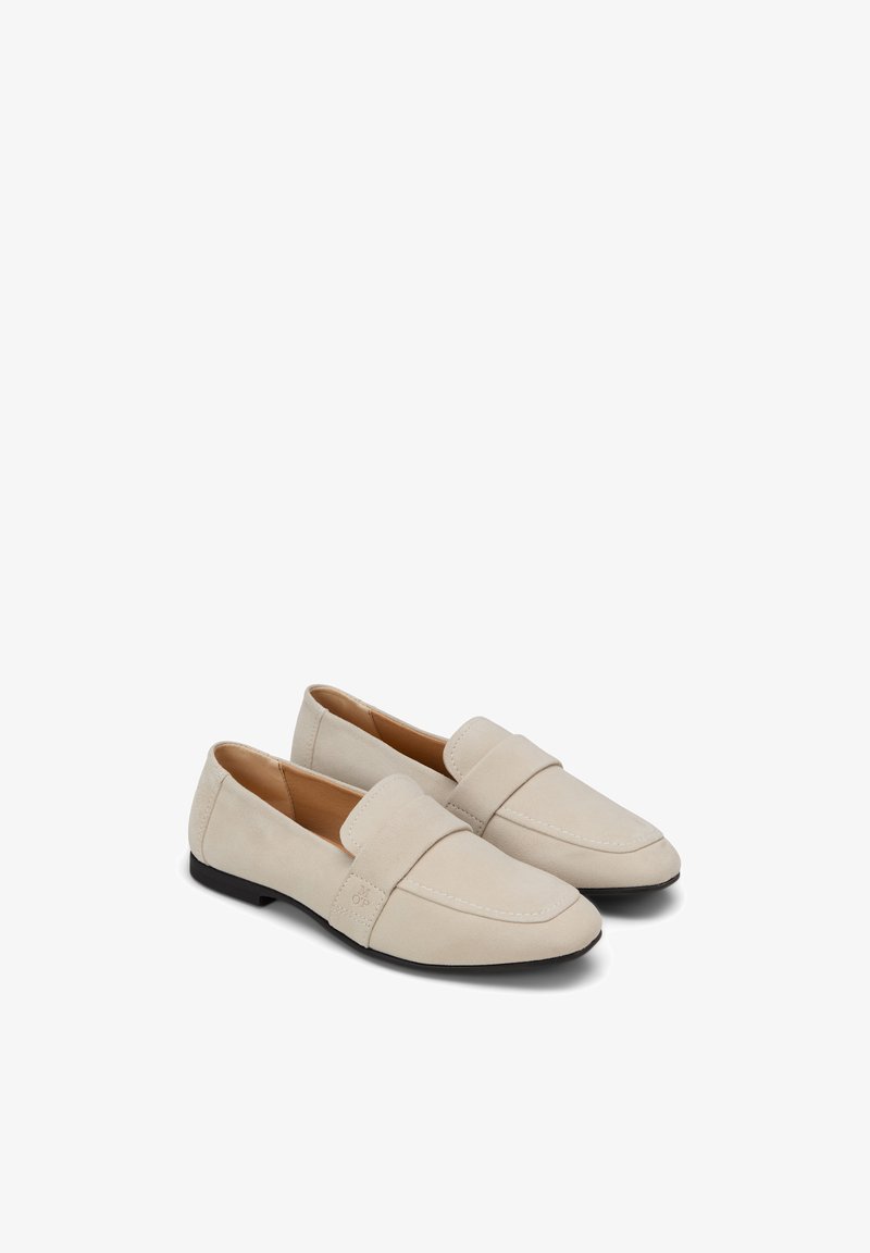 Paire de mocassins beige à enfiler, avec des bouts ronds, des talons bas noirs et une large lanière sur le dessus, présentés sur un fond blanc.