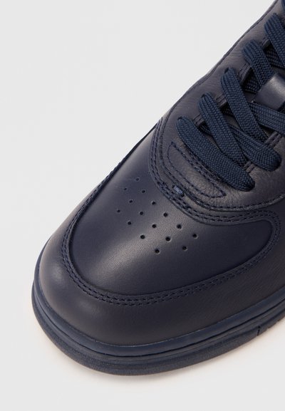 Mornarsko modri usnjeni superge s perforiranim nosom, teksturiranimi vezalkami in kontrastnim šivanjem. Zaobljen nos in debel gumijast podplat sta vidna.