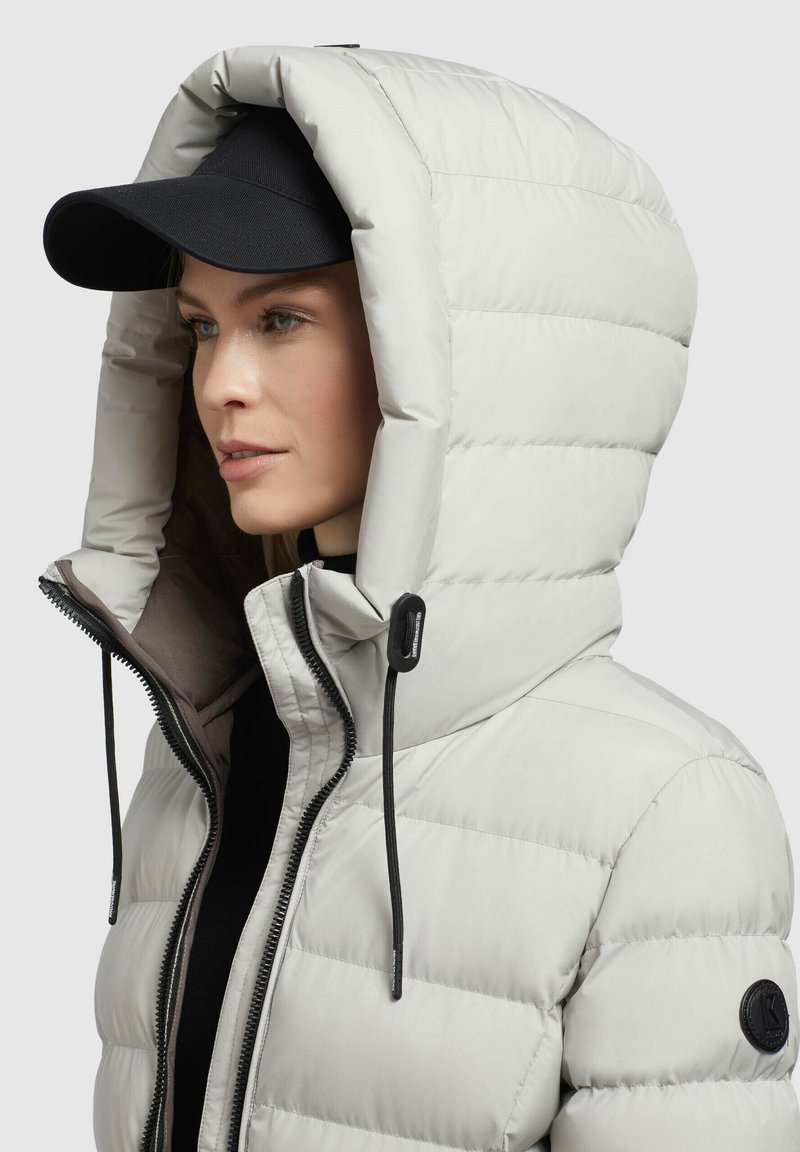 Hellgraue Daunenjacke mit großer Kapuze, Reißverschluss, elastischen Zugbändern und einem schwarzen Stoff-Logopatch am Ärmel.