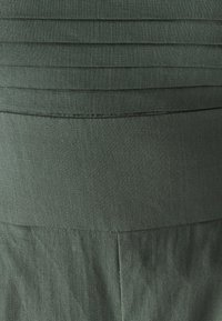 Donkergroene rok met geplooide textuur aan de bovenzijde en gladde stof eronder, met een gestructureerde vorm en zichtbare naden.