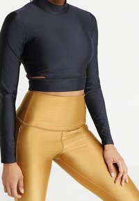 Navy-blaues langärmeliges Crop-Top mit hohem Kragen und seitlichen Ausschnitten, kombiniert mit hochgeschnittenen, goldenen Leggings mit glatter, glänzender Textur.
