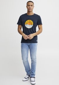 Marine T-shirt med et rundt grafik, der viser solen, der stiger op over stiliserede bølger, parret med lyseblå jeans og marine- og hvide sneakers.