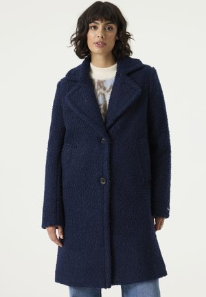 Woman with short dark hair wears a textured navy blue coat over a patterned sweater and blue jeans, standing against a plain light background.