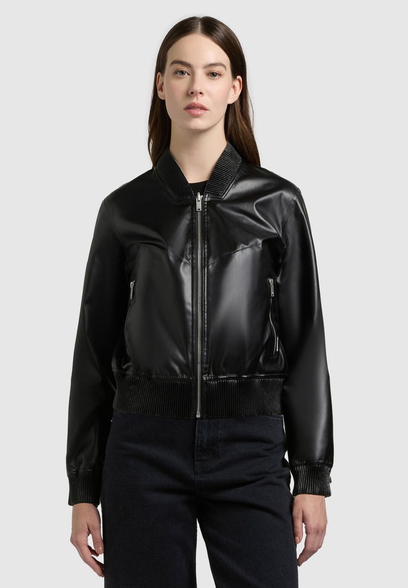 Jeune femme portant une veste bomber noire en similicuir à fermeture éclair, avec col, poignets et ourlet côtelés, debout devant un fond clair uni.