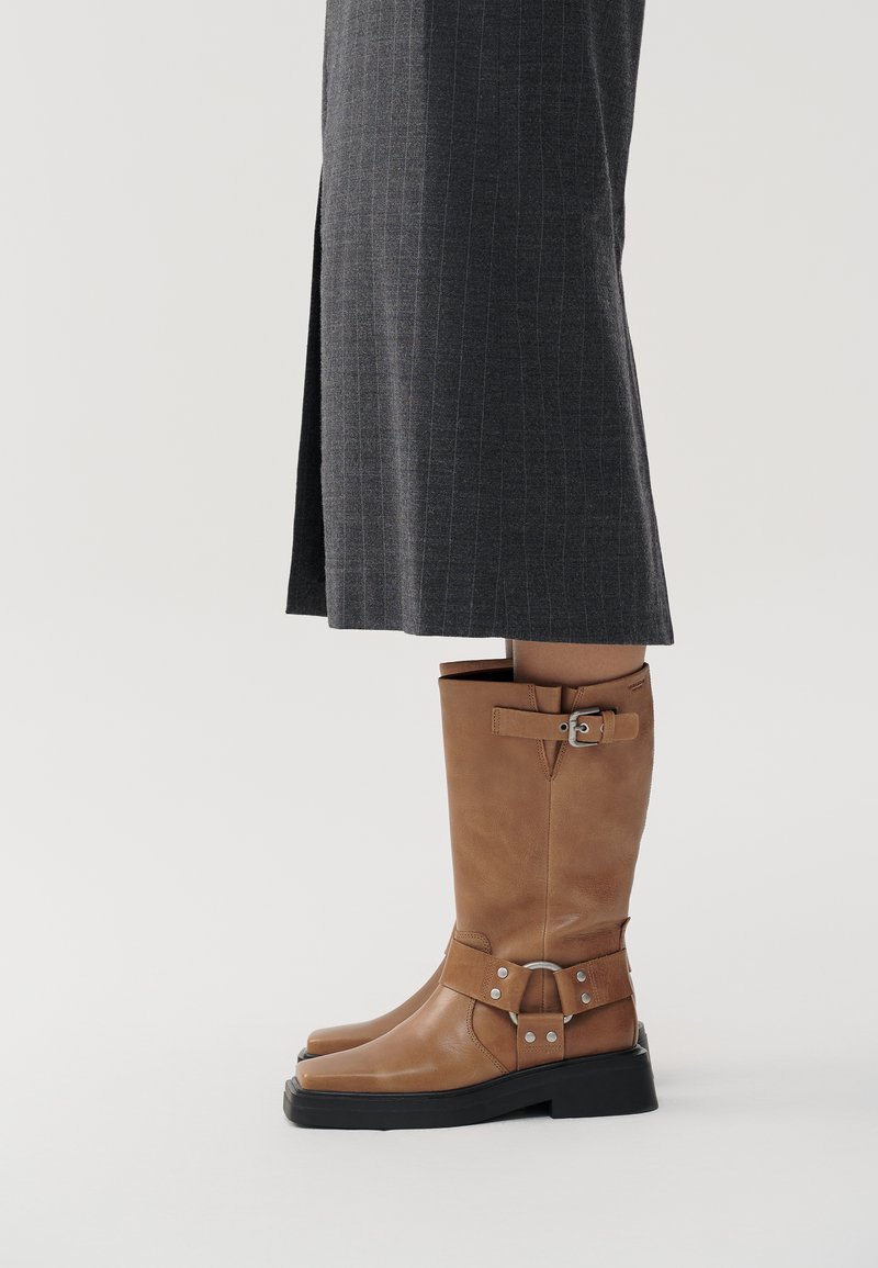 Botas de cuero marrón hasta la rodilla con suela plana, detalle de correa ajustable con herrajes plateados y textura suave. Suela de goma negra.