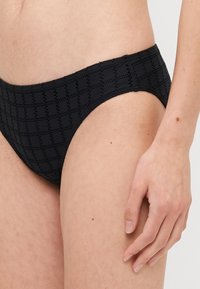 Svart bikinitrosa med ett texturerat rutmönster, midjehögt snitt och släta kanter. Tyget verkar stretcha och vara lätt.