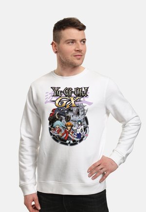 Mężczyzna w białej bluzie z kolorowymi postaciami z anime Yu-Gi-Oh! GX i logo, stoi z jedną ręką na biodrze, patrząc na bok.