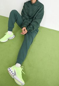 Osoba ve tmavě zelené teplákové soupravě sedí na světle zeleném koberci, má na nohou neonově žluté tenisky a bílé ponožky, ruce sepjaté před sebou.