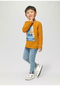Langmouw shirt in mosterdkleur met grafisch ontwerp, blauwe denim jeans en witte sneakers met grijze accenten en zwarte buitenzool.