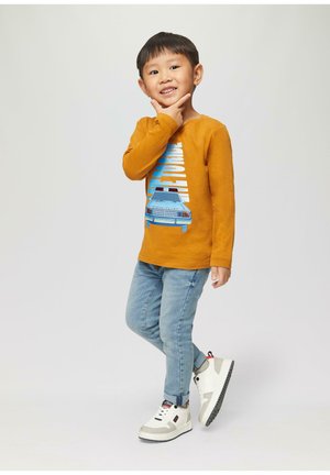 Chemise à manches longues couleur moutarde avec un design graphique, jean en denim bleu et baskets blanches avec des accents gris et une semelle extérieure noire.