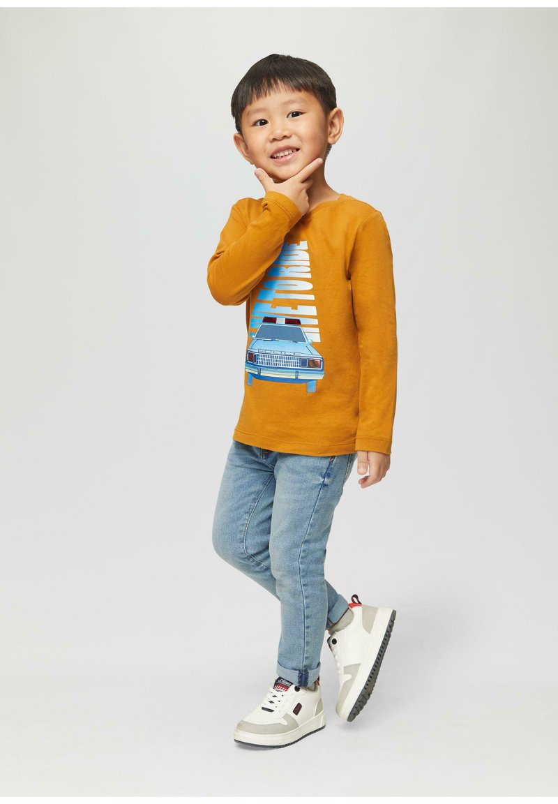 Langmouw shirt in mosterdkleur met grafisch ontwerp, blauwe denim jeans en witte sneakers met grijze accenten en zwarte buitenzool.