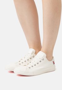 Bílé plátěné tenisky s pěti otvory pro tkaničky, vyšívaným květinovým detailem na vnější straně a texturovanou gumovou podrážkou. Nízký střih s tkaničkami.