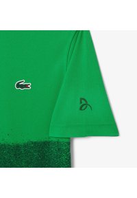 Grön polo skjorta med en texturerad tygkvalitet, med Lacoste krokodil-logotypen och en svart detalj på vänster ärm. Gradvis mörkgrön effekt i botten.