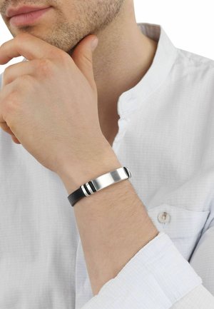 Hombre con camisa blanca apoyando la mano en la barbilla, mostrando una pulsera negra de cuero con placa metálica plateada en la muñeca.