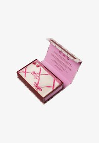 Silke sovemaske i en kremfarge, plassert i en rosa og rød boks. Boksen har mønstrede bånd og inneholder produktinformasjon inni.