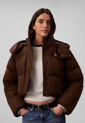 Kvinde iført en brun dunjakke over en hvid cropped sweater og blå jeans med hænderne i lommerne mod en grå baggrund.