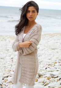 Mujer con largo cabello oscuro que lleva un cárdigan beige de punto y un conjunto blanco, de pie en una playa rocosa con el mar de fondo.