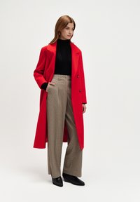 Czerwony wełniany płaszcz z podwójnymi guzikami, czarny prążkowany golf i spodnie w kratę z wysokim stanem. Czarne botki na obcasie dopełniają całość.