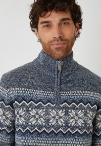 Gebreide trui in marineblauw met sneeuwvlok- en zigzagpatronen in wit en lichtblauw. Heeft een hoge kraag en een ritssluiting. Zachte textuur.