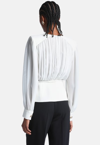 Femme aux cheveux tressés vue de dos, portant un chemisier blanc plissé à manches longues et un pantalon noir sur un fond uni.