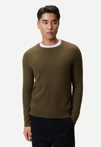 Olivgrön ribbad tröja med rund halsringning, långa ärmar och en texturerad stickning. Bärs över en vit T-shirt, kombinerad med mörka byxor.