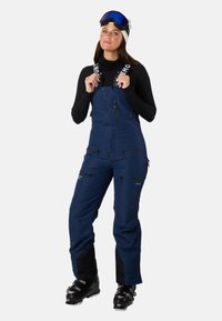 Navy blue ski overalls with adjustable straps, multiple zippered pockets, and black accents at the ankles, made from durable weather-resistant material.