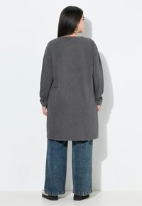 Túnica gris de manga larga con un corte relajado, que presenta un cuello redondo y aberturas en los laterales, combinada con unos pantalones vaqueros de mezclilla azul de pierna ancha.
