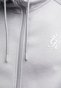 Felpa con cappuccio grigio chiaro con chiusura lampo e vestibilità aderente, caratterizzata da una texture morbida, coulisse regolabili e un logo a forma di corona bianco sul petto.