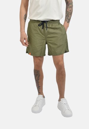 Uomo che indossa pantaloncini verde oliva con coulisse, scarpe da ginnastica bianche e camicia bianca, mostrando tatuaggi su braccia e gamba sinistra.