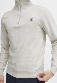 Light beige sweatshirt featuring a quarter-zip design, ribbed cuffs and hem, and a small black logo on the chest. Smooth cotton material.