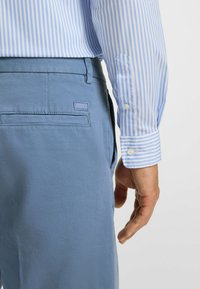 Light blue pants with a textured fabric and a logo patch, paired with a blue and white striped shirt with buttoned cuffs.