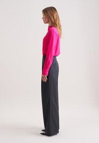 Fuchsia blouse met lange mouwen, gecombineerd met donkergrijze comfortabele pantalon met een subtiele streep. Model draagt puntige zwarte hakken.