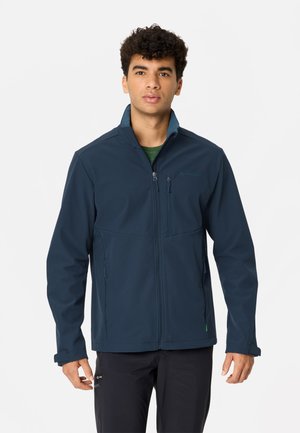 Junger Mann mit lockigen Haaren, der eine dunkelblaue Zip-Jacke und schwarze Hose trägt, steht vor einem schlichten hellen Hintergrund.