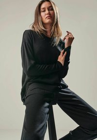 Black knit sweater with relaxed fit and ribbed cuffs, paired with matching black tailored pants. Model poses on a stool, showcasing the outfit.