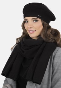 Černý baret a šála vyrobená z jemné tkaniny. Šála se omotává kolem krku. Modelka má na sobě šedý kabát přes černý top.