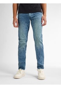 Jean azul de mezclilla con un lavado desgastado, corte ajustado y ligero estiramiento. Presenta bolsillos frontales, cremallera y zapatillas blancas.