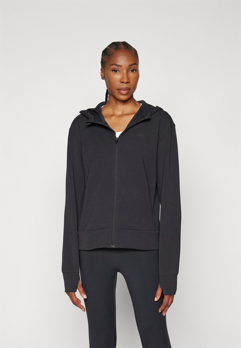 Black zip-up hoodie with a hood, made of soft fabric. Features long sleeves and a fitted waist, minimal branding on the chest.