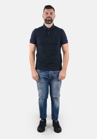 Polo shirt navy, maniche corte, tessuto strutturato. Indossato con jeans blu sbiaditi e scarpe da ginnastica nere. L'uomo sta in piedi, rivolto in avanti.