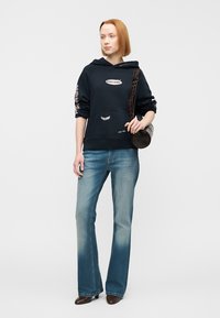 Σκούρα μπλε φούτερ με κουκούλα, μπροστινή τσέπη, λευκό λογότυπο και λουλουδάτες λεπτομέρειες στα μανίκια, συνδυασμένο με ανοιχτό μπλε τζιν καμπάνα και στρογγυλή τσάντα χιαστί.