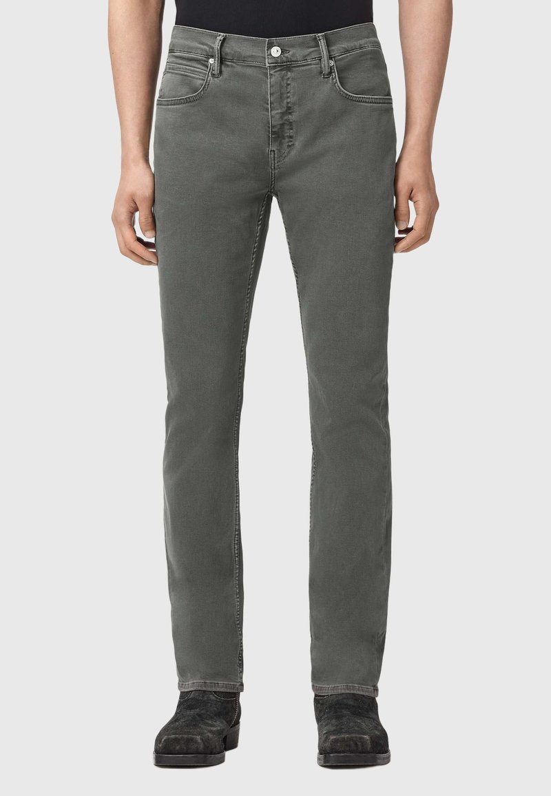 Mand iført mørkegrå slim-fit jeans, sorte støvler og en sort top, stående mod en ensfarvet lys grå baggrund.