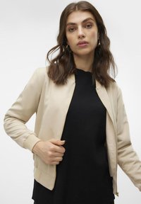 Beige Bomberjacke mit glatter Textur, Reißverschluss vorne, gerippten Bündchen und Saum. Getragen mit einem schwarzen Pullover darunter.