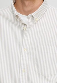 Camicia a righe di colore crema chiaro con sottili strisce grigie, dotata di colletto classico e taschino sul petto sinistro, con una texture di tessuto naturale.