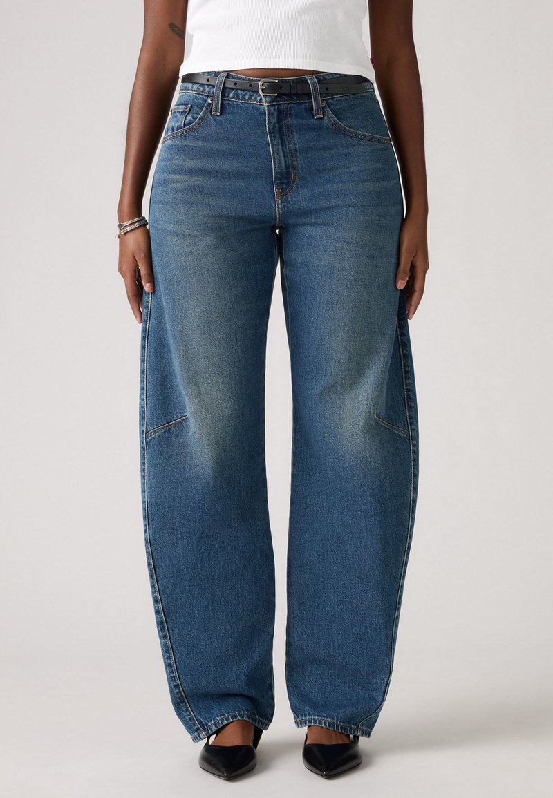 Jean bleu large en denim, avec une taille haute, des poches avant et une légère décoloration. Stylé avec une ceinture noire et des chaussures à bout pointu.