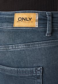 Jeans de mezclilla de un tono azul oscuro, con una etiqueta rectangular en color beige que lleva el texto "ONLY", y costuras visibles a lo largo de los bordes y bolsillos.