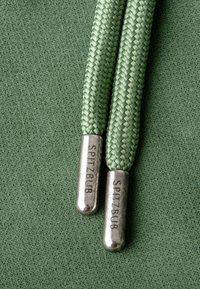 Lacci intrecciati verdi con punte metalliche argentate marcate "SPITZBUB" poggiati su tessuto verde, che mostrano contrasto di texture e colore.