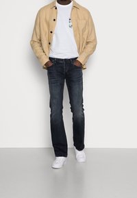 Mężczyzna ubrany w beżową koszulę zapinaną na guziki, na t-shircie z kwiatową kieszonką, ciemnoniebieskie dżinsy, białe trampki, z rękami w kieszeniach, stoi na jednolitej podłodze.