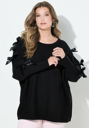 Mujer con cabello largo y ondulado que lleva un suéter negro decorado con lazos de satén en las mangas y pantalones rosa claro, de pie contra una pared blanca.