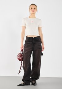 Witte geribde top met korte mouwen en rode hartborduursels, gecombineerd met losse zwarte denim broeken en een kleine bordeauxrode handtas. Zwarte puntige schoenen.